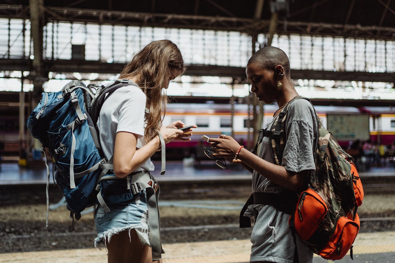 Two young women travelers using smartphones at a train station, planning their journey.